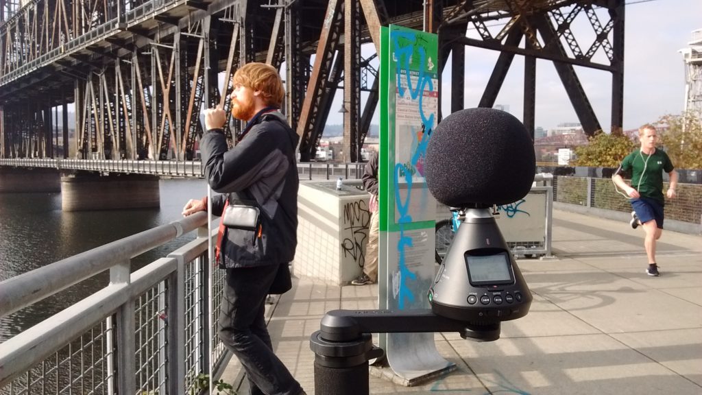 In this photo, an ambisonic recorder in the foreground records spatial audio, a man is jogging to the right of and a man stands looking at the Willamette River while recording binaural audio sounds. The Steel Bridge is in the background. Photo by Mary Anne Funk. 